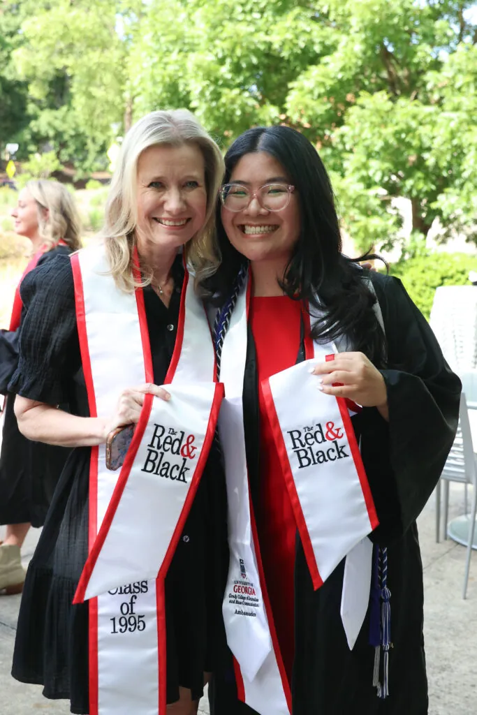 Two women smile and stand close together outdoors, wearing black graduation robes and white sashes with red trim that read Red & Black. One sash says Class of 1995. There are trees and people in the background.