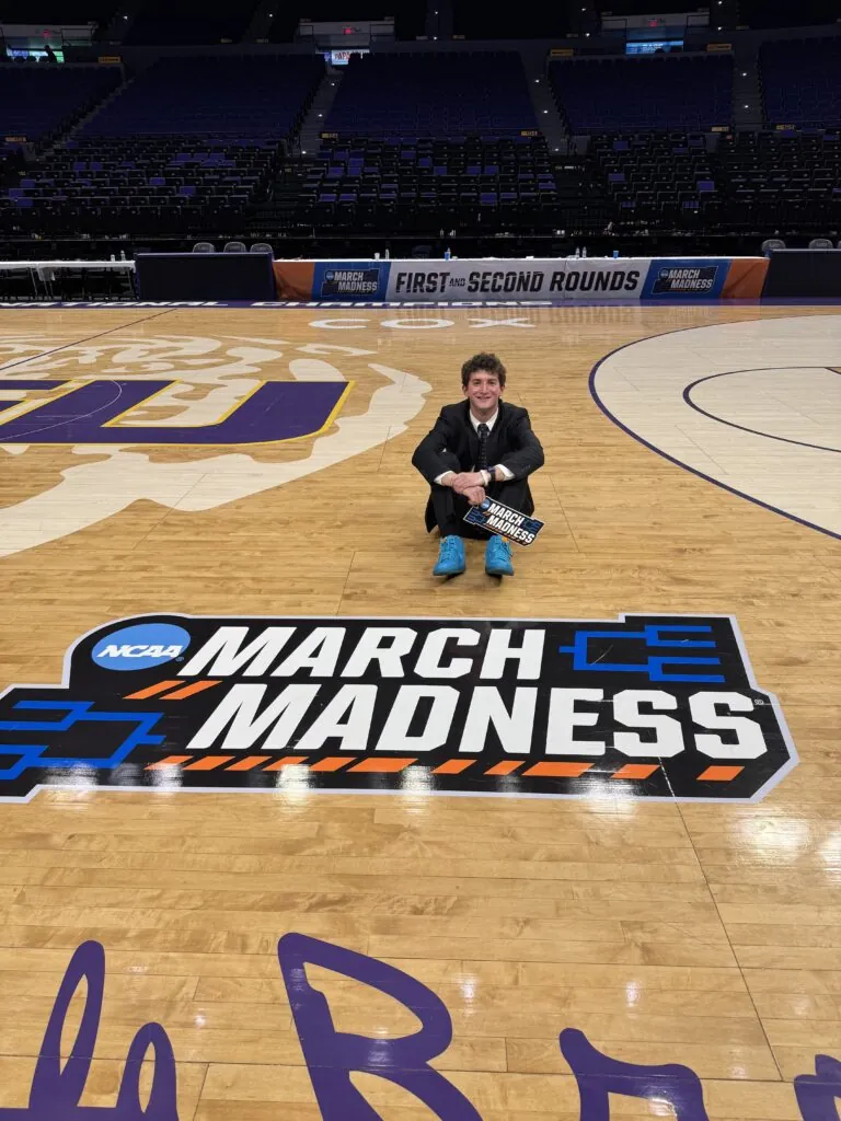 A person in a suit with teal shoes sits cross-legged on a basketball court, holding a sign, behind a large NCAA March Madness logo. Empty arena seats and March Madness banners are visible in the background.