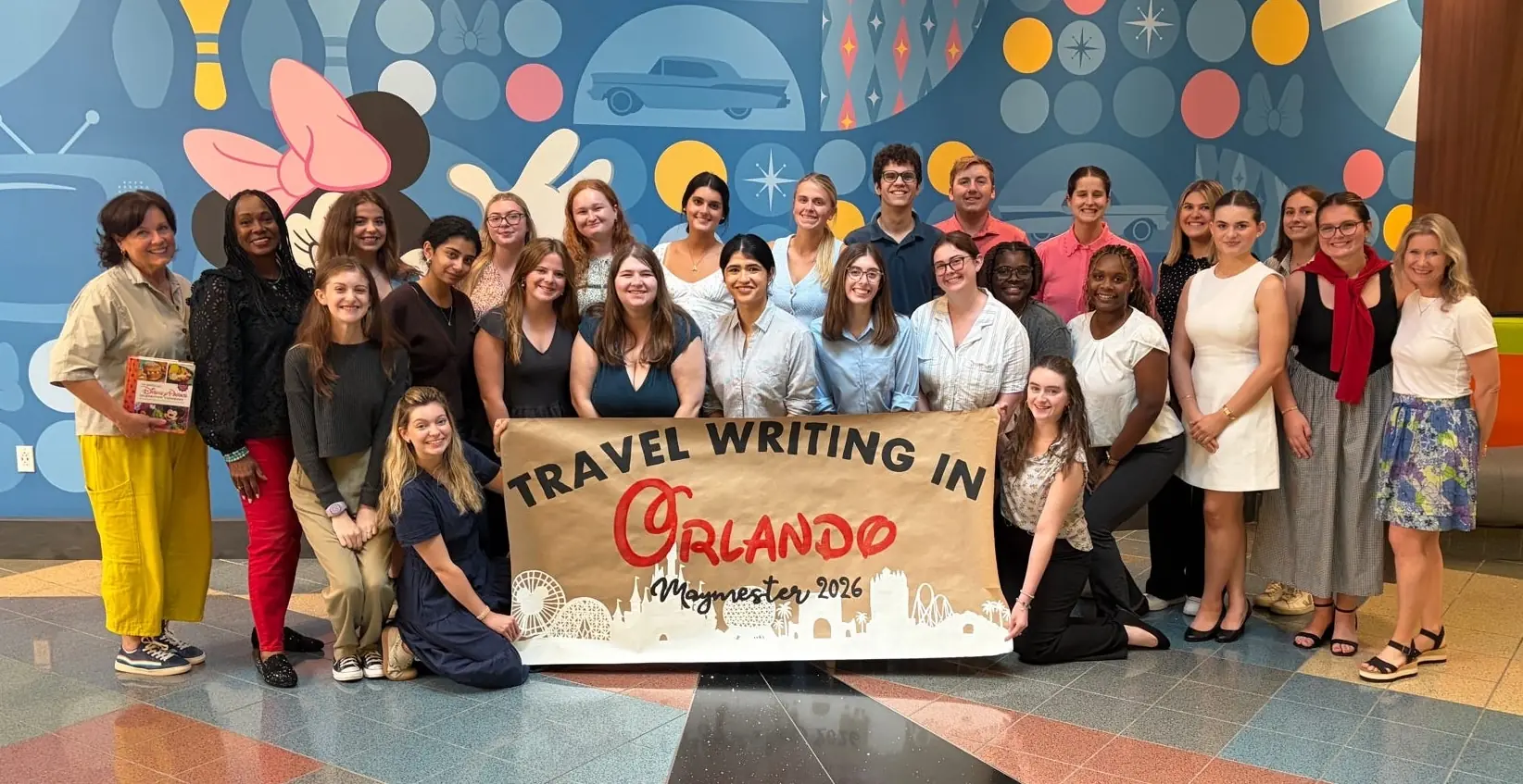 A large group holds up a banner that reads "travel writing in Orlando Maymester 2026."