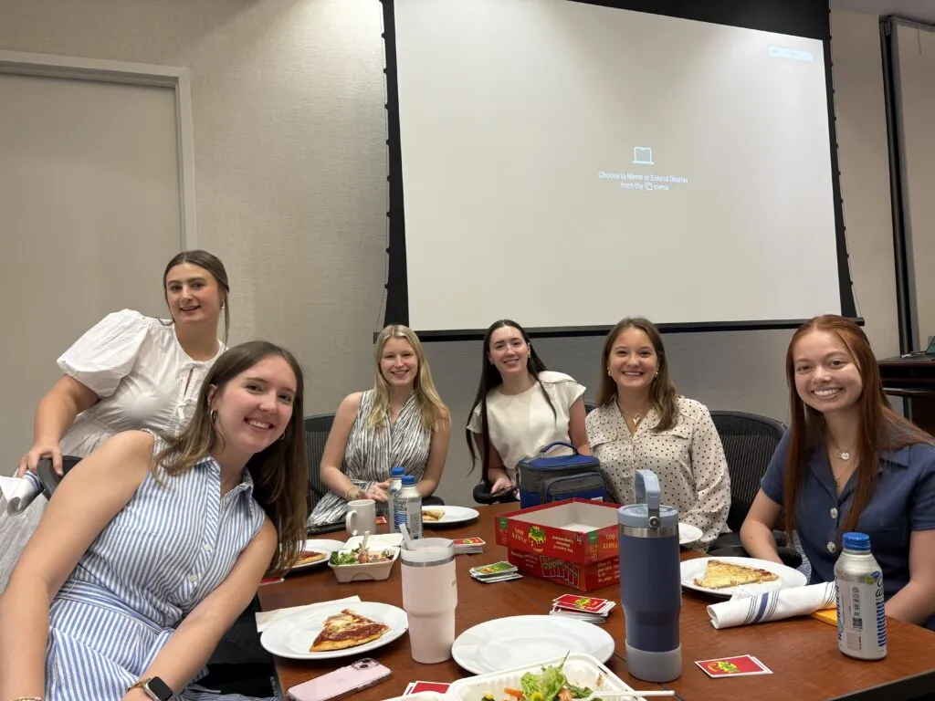 Six young women sit around a table with pizza, salad, drinks, and Uno cards, smiling at the camera in a conference room with a blank projector screen in the background.