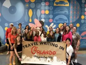 A large group holds up a banner that reads "travel writing in Orlando Maymester 2026."