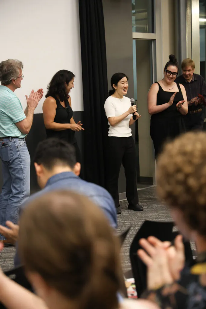 Susan Noh talks into a microphone as other faculty around her applaud.