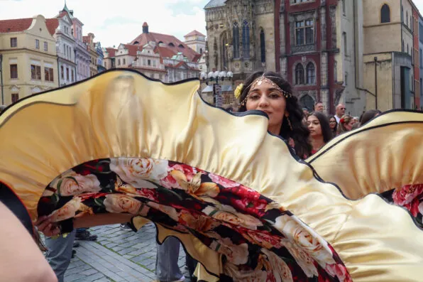 A woman in a decorated dress with yellow ruffles and floral patterns dances in a historic European square, capturing the vibrant essence of Travel Writing Prague, surrounded by people and old buildings.