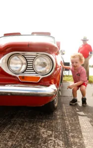 A toddler smiles as he looks at his reflection in a red car at the fair.