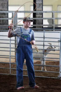 A teenager demonstrates a lasso while a goat looks on in the background at the fair.