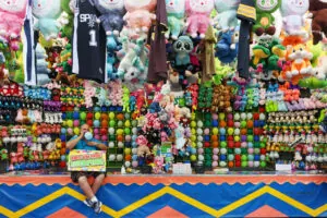 A colorful scene with balloons and stuffed animals and a man sitting blowing up a ballon at a carnival attraction at the fair.