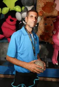 A man in a blue shirt holding a ball looks serious as he stands in front of a display of stuffed animals at the fair.