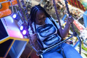 A girl wearing a shirt with a bunch of mouths on it smiles as she swings on a ride at the fair.