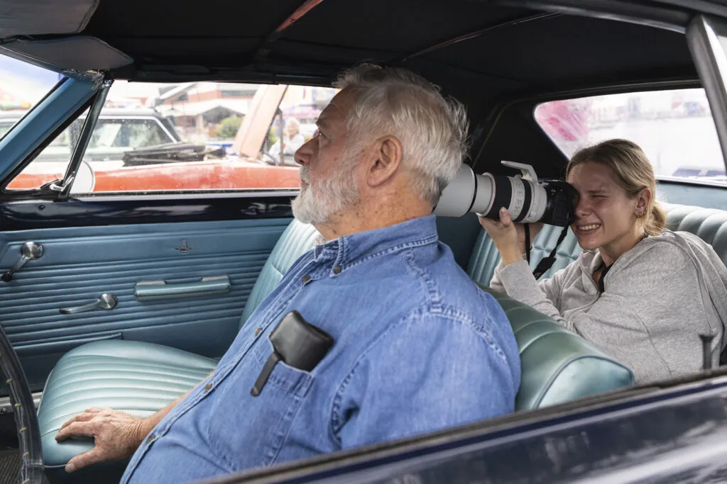 A student with a camera sits in the backset of a car and photographs a man through his rear view mirrow.