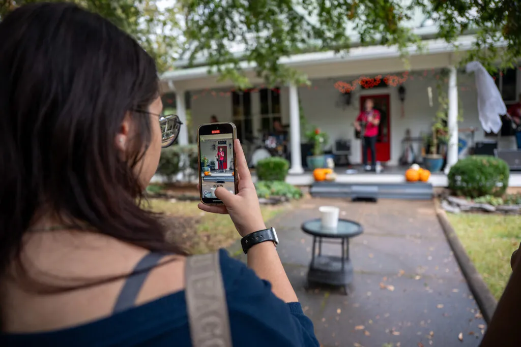 The camera looks over the shoulder of a student filming a band on a front porch.