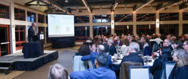 Charles Davis stands at a lectern and addresses a group of people at tables.