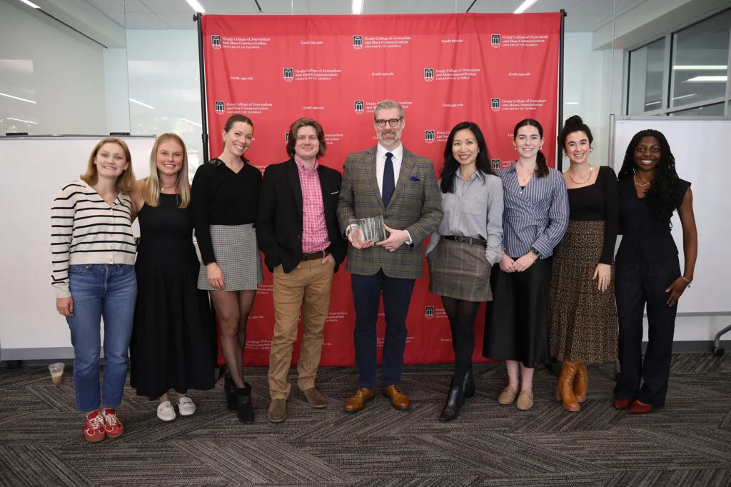 Simon Paterson holds his trophy while surrounded by a group of faculty and students in front of a Grady College step and repeat.
