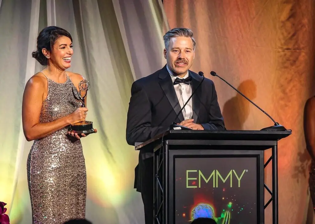 Laura Evans holds an Emmy Award as Tamlin Hall speaks at a podium with the word "Emmy" written on it.