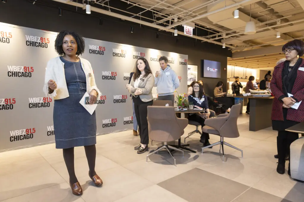 Tracy Brown speaks to a small group inside a modern office space with WBEZ 91.5 Chicago logos on the wall. Some people stand while others sit at tables, listening attentively.