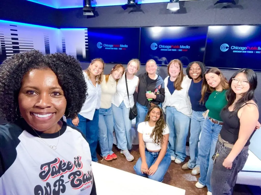 A group of twelve people, mostly young women, stand together smiling inside a radio studio with Chicago Public Media logos on the walls. Tracy Brown stands in front, taking a selfie with the group gathered happily in the background.