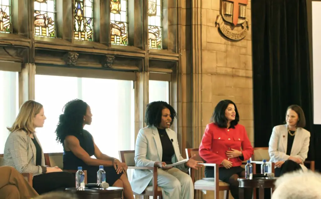 Five women, including Tracy Brown, sit on a panel discussion in front of stained glass windows and a stone wall with a crest, each dressed in business attire and engaging in conversation with water bottles on the table.
