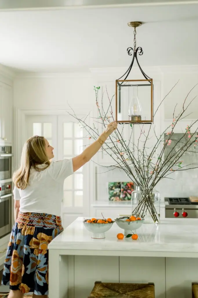 Robin Burnett is pictured in a space designed by her company, Robin Burnett Design. She is reaching up to straighten a lamp above an island in a kitchen.