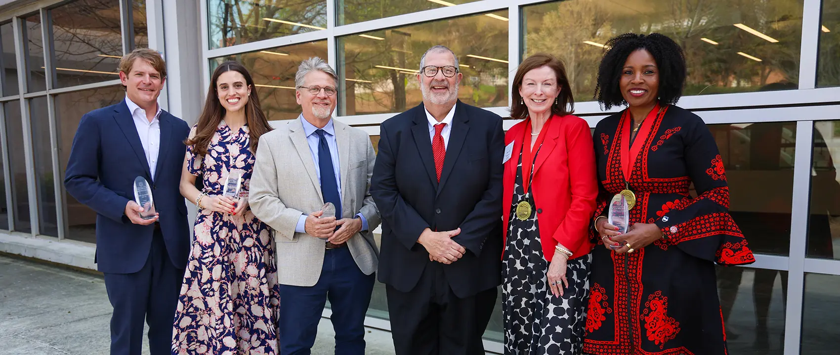 A group of alumni holding awards and wearing medals stand with Dean Davis in front of Grady College.