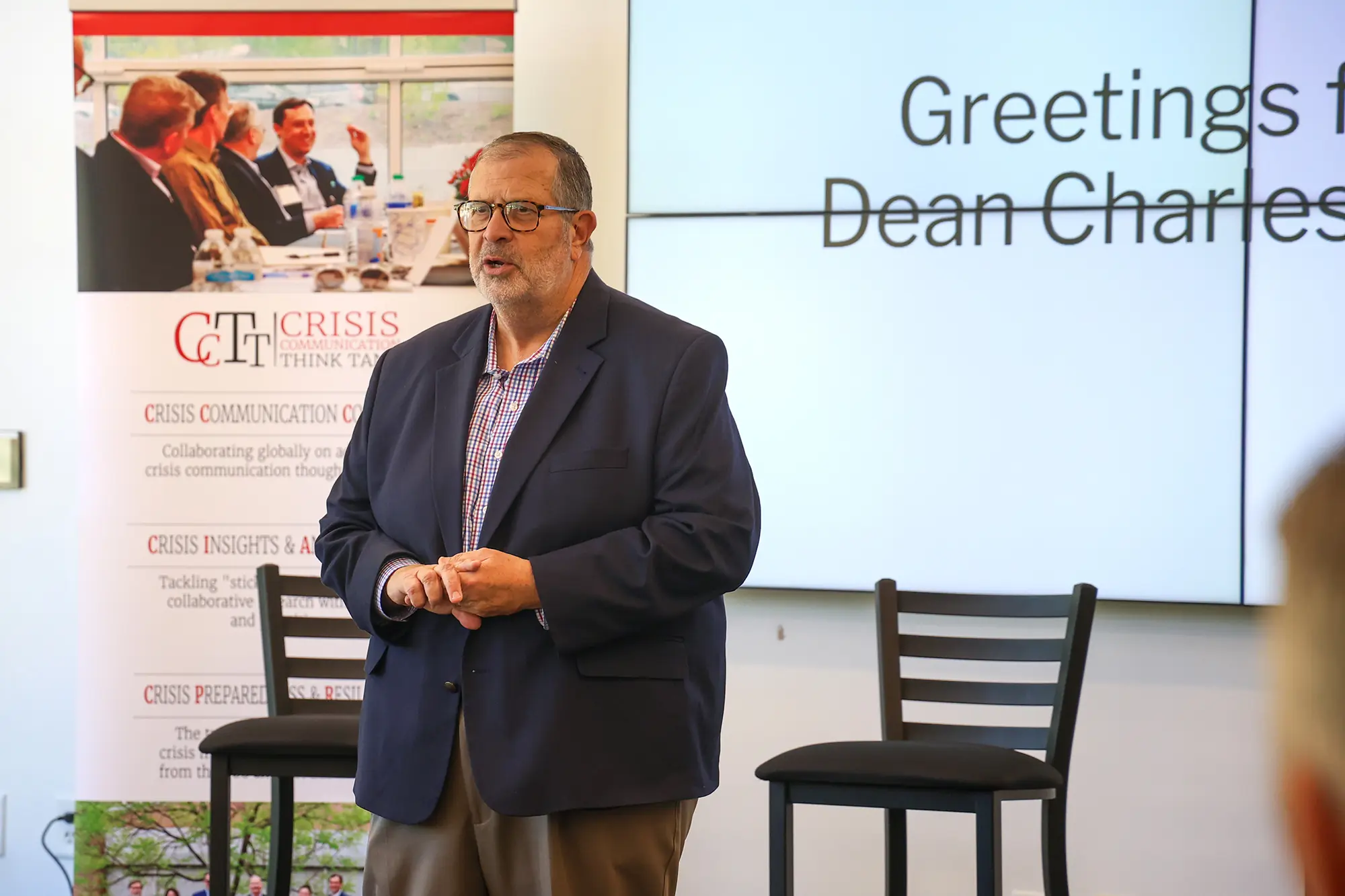 A close-up of Dean Charles Davis speaking in front of a group. He is a man with glasses and a beard, wearing a navy blue blazer over a checkered shirt. The background shows a blurred digital screen with the text "Greetings from Dean Charles..." and a CCTT banner to the left.