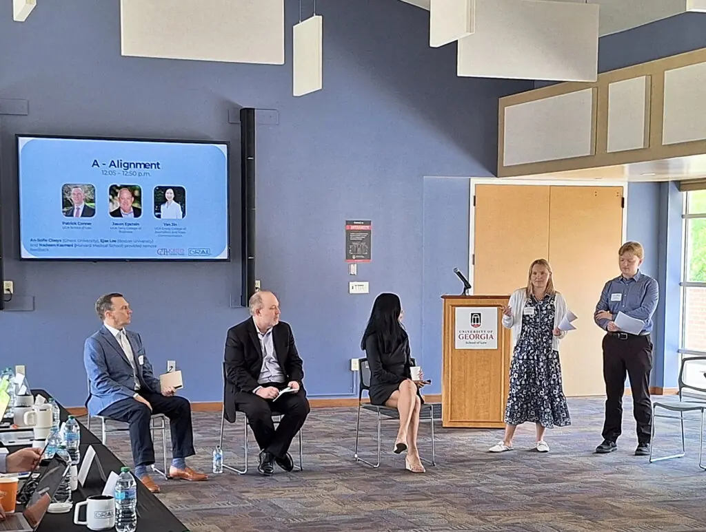 Three people sit and two people stand as they present in front of a digital sign that reads A - Alignment