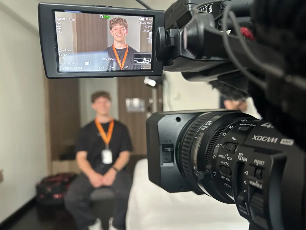 Landon Shell, a young man with a lanyard, sits for an interview in a room, smiling at the camera. The scene is filmed by a professional video camera, which captures Landon’s image on its display screen in the foreground.