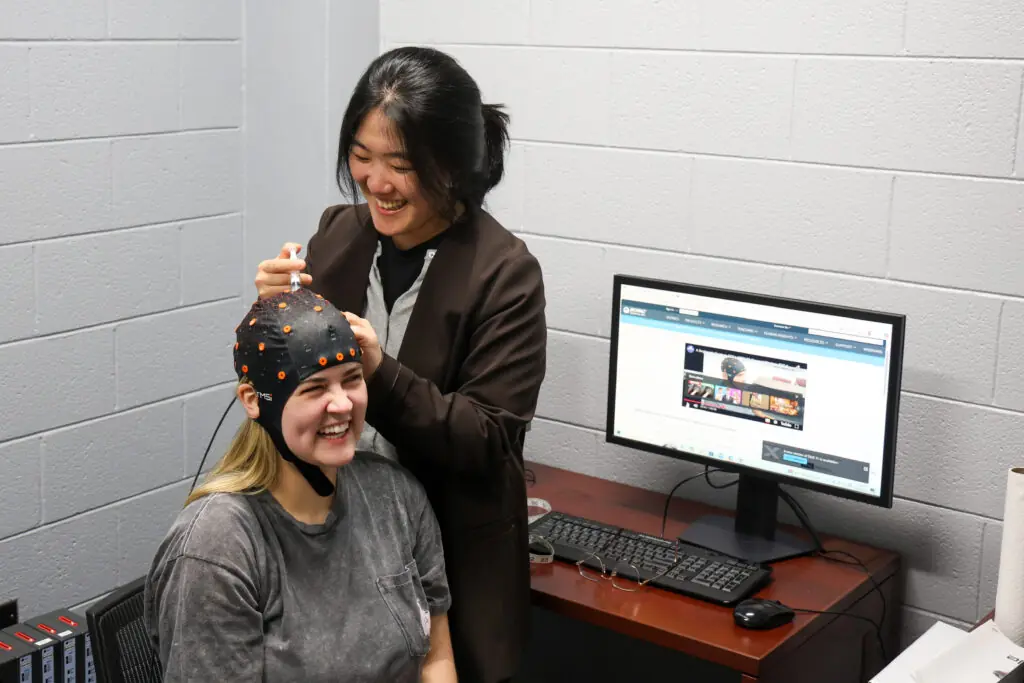 Two women in a neuroscience lab; one smiles, wearing a sensor cap as Brittany Shivers adjusts it. Behind them, a computer displays research websites, highlighting their focus on brain science and innovation.