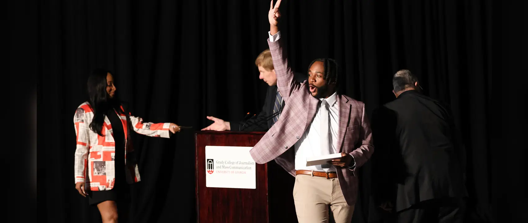 A man gives an YO hand sign for his fraternity Kappa Alpha Psi as he crosses a stage. He is holding a box in his hand and there is a lectern in the background reading Grady College of Journalism.