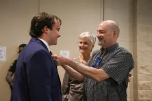 A candid moment where an older man in a patterned shirt adjusts the tie of a younger man in a navy blue suit. An older woman with short grey hair and glasses stands between them, watching with a warm smile.