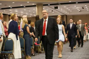A man in a black suit and red tie leads a procession of graduates and faculty down the center aisle of a large event hall. The audience stands on either side, watching as they walk toward the front.