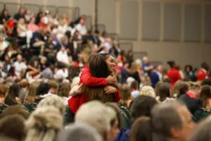 A high-angle, candid shot of a graduate in a red blazer embracing another person in the middle of a large auditorium. The background is filled with a soft-focus audience seated in bleachers and chairs.