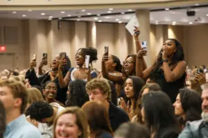 A lively group of young women in an auditorium cheering and holding up their smartphones to take photos or videos. Their expressions are joyful, with some laughing and shouting in celebration.