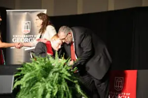 On a graduation stage, an older man in a suit leans down to warmly embrace a graduate in a wheelchair. A woman in a white patterned blazer stands in the background, smiling as she shakes hands with another person off-camera.
