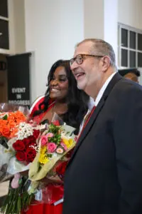 A close-up profile shot of a smiling graduate with long dark hair holding several large, vibrant bouquets of roses and daisies. She stands next to a smiling older man in a suit and glasses.