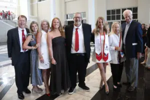 A group of eight family members and graduates pose for a multi-generational photo in a modern, bright lobby with marble floors. The group includes two graduates in white dresses and red stoles, surrounded by smiling adults in formal and semi-formal attire.