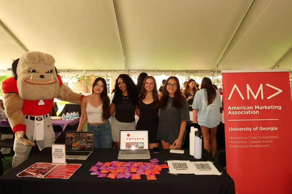 Five people, including Sophia Kim and a mascot in a bulldog costume, stand smiling behind a table with marketing materials and candy. A red American Marketing Association sign for the University of Georgia is displayed on the right.