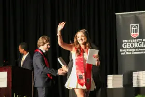 A female graduate in a short white dress and a red University of Georgia stole smiles and waves to an audience while walking across a stage. She holds a diploma cover in her left hand. Two men in suits stand in the background behind a podium.