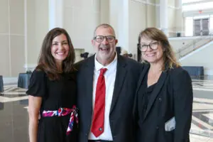 Three adults pose for a photo in a brightly lit lobby. A man in a suit and red tie stands in the center with his arms around two women, all three smiling broadly. The woman on the left wears a black dress with a colorful sash, and the woman on the right wears a black blazer.