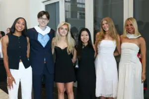 Six young adults stand in a row posing for a group photo in a modern hall. They are dressed in semi-formal attire, including black and white dresses and a blue suit. Two of the individuals wear white academic hoods over their shoulders.