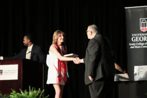 A female graduate with blonde hair shakes hands with a man in a suit on a stage. She wears a white dress and a red stole with teal tassels. A large banner for the University of Georgia Grady College is visible to the right.