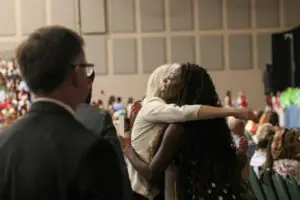 A candid, over-the-shoulder shot of a woman with long, dark curly hair hugging another person in a white jacket. They are in a large indoor venue filled with people seated in green chairs during a ceremony.