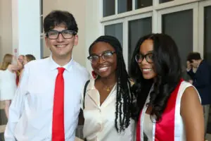 Three young adults smile for a close-up photo. The man on the left wears a white shirt and bright red tie; the two women beside him wear white tops and red academic stoles. They are all wearing glasses.