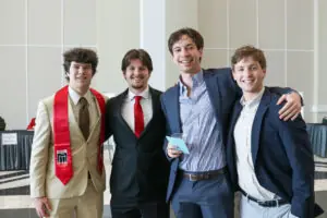 our young men in suits and dress shirts stand with their arms around each other, smiling for a photo in a large hall. One man on the far left wears a tan suit and a red University of Georgia stole.