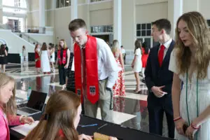 A high-angle shot of a check-in desk at an event. Two women are seated at the table with a laptop and paperwork, while several students, some wearing red graduation stoles, stand in line to check in.
