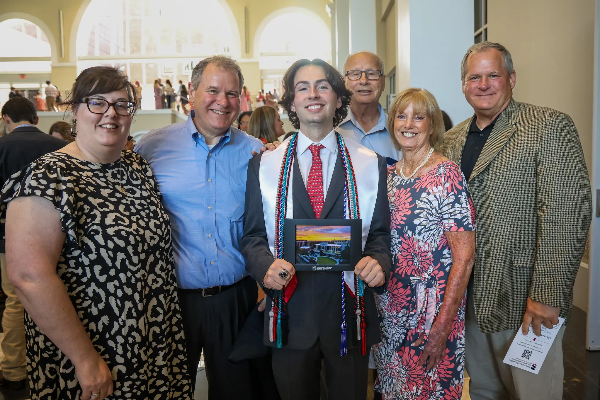 A male student holds his framed photo of Grady College and is surrounded by smiling family members with large atrium windows behind.