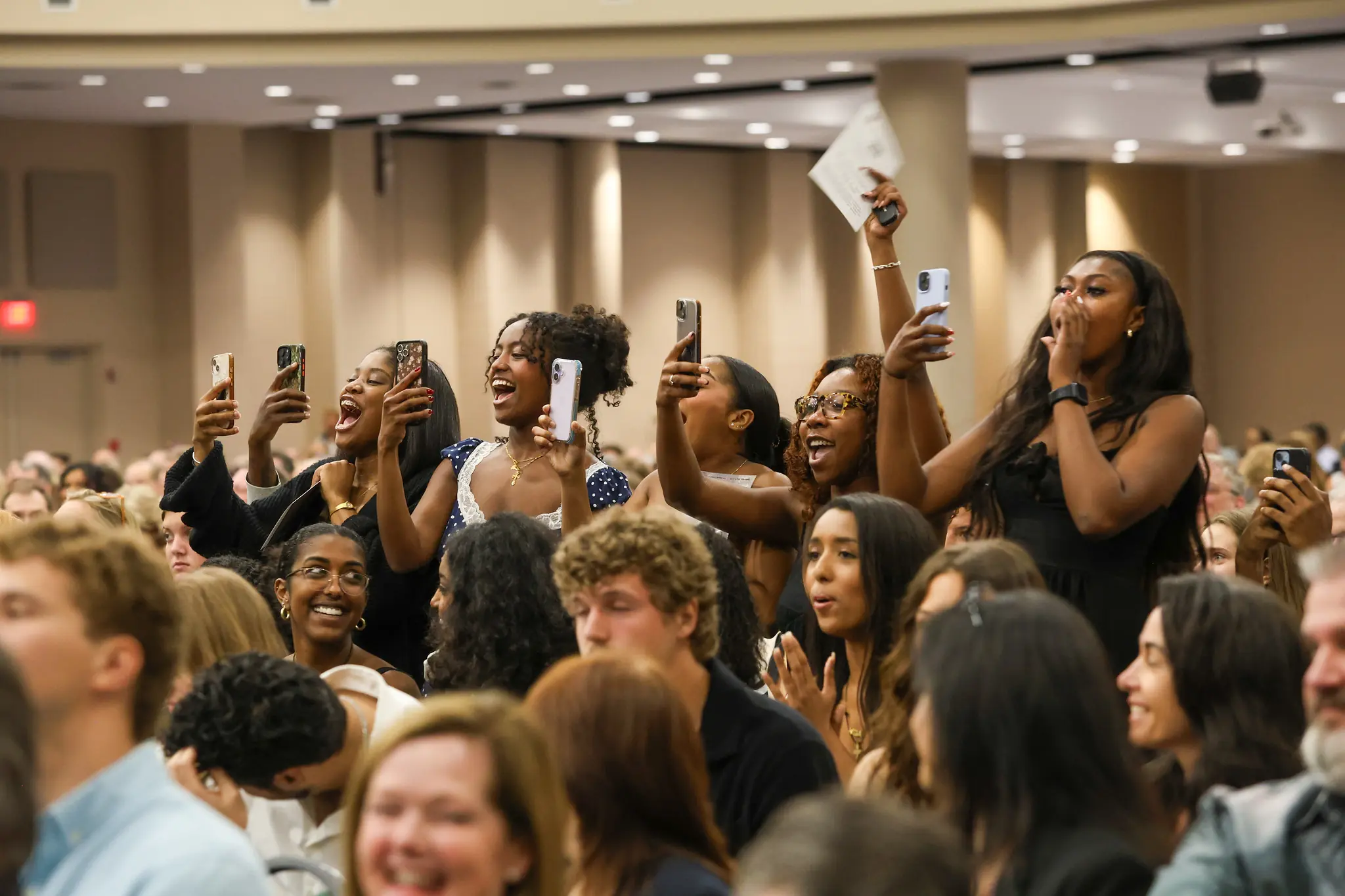 Several people stand up in a crowd and cheer as they hold phones to take pictures.