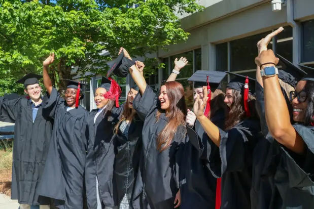 A side, close-up view of about 8 graduates wearing graduation caps and gowns cheering outside of Grady College.