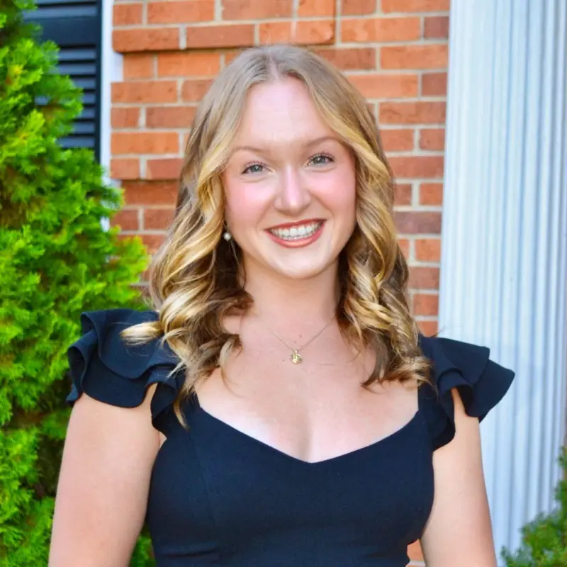 Bethany Barner, a woman with brown, curled hair smiles at the camera. She is wearing a navy top and standing against a brick background.