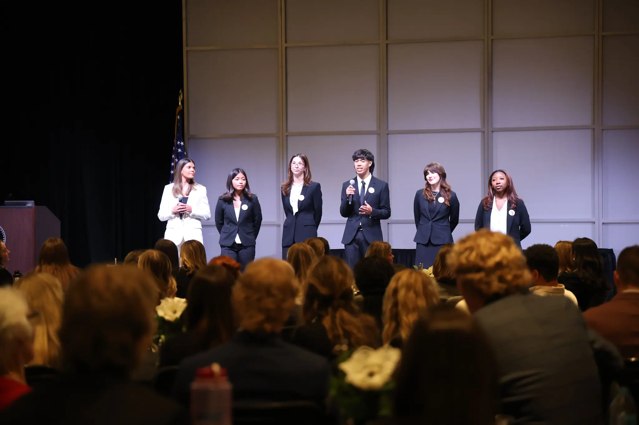 Six students dressed in business attire make a presentation. The audience is shown in the foreground.