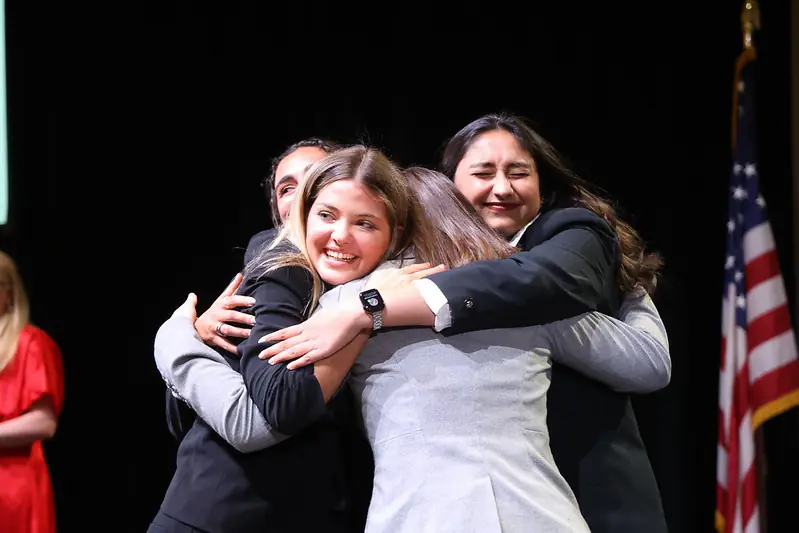 Four people in business attire share a group hug on stage, smiling and celebrating. An American flag is visible in the background.