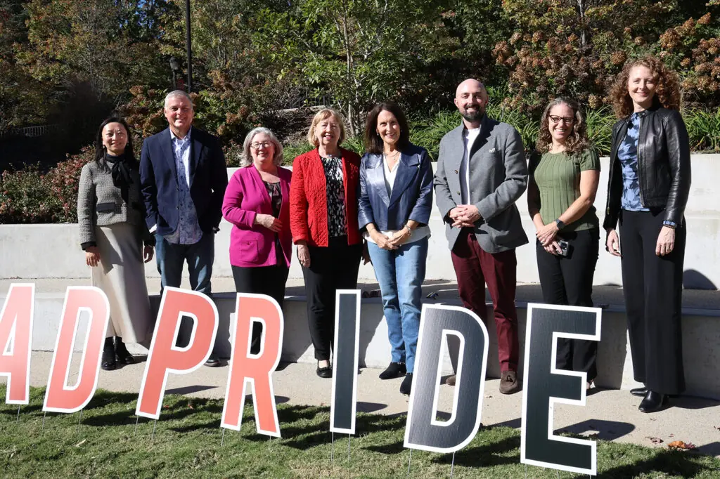 Eight people stand outdoors behind large letters spelling “PRIDE,” smiling at the camera with trees and greenery in the background.